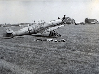 Piloot ligt in het gras naast zijn jager te slapen. Op de achtergrond een boederij aan de Schipholweg 115.